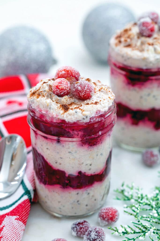 View of cranberry overnight oats in jar with second jar, silver ornaments, spoon, and sugared cranberries in background