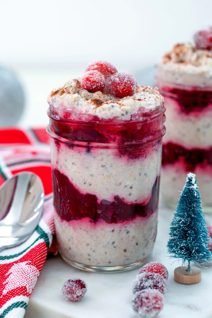 Head-on view of a small jar of cranberry overnight oats topped with sugared cranberries with small glitter Christmas tree, second jar of oats, and spoon
