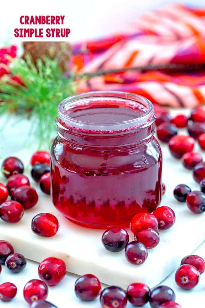 Small jar of cranberry simple syrup with fresh cranberries all around and recipe title at top.