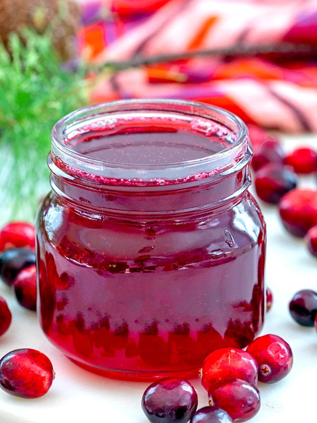 Close-up view of a small jar of cranberry simple syrup with fresh cranberries all around and recipe title at top