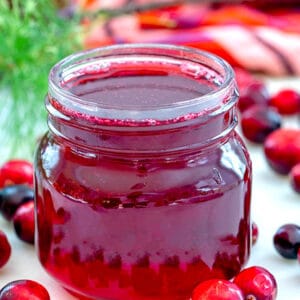 Close-up view of a small jar of cranberry simple syrup with fresh cranberries all around and recipe title at top