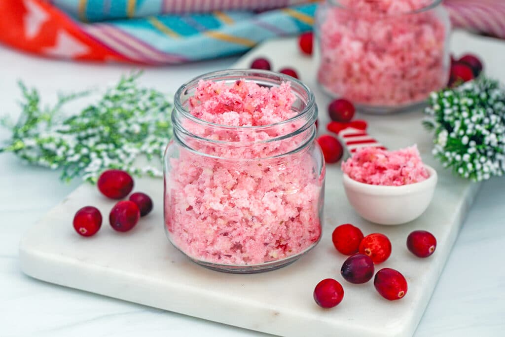 Head-on landscape view of a small mason jar of cranberry sugar with second jar and spoonful of sugar in background and fresh cranberries all around