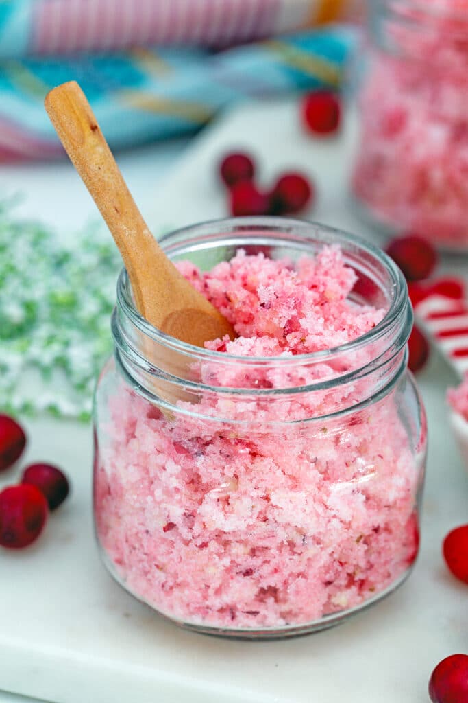 Small mason jar of pink cranberry sugar with small wooden spoon and fresh cranberries all around