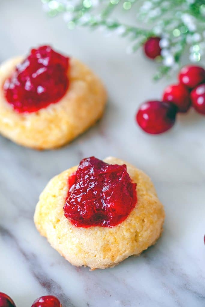 View of two cranberry thumbprint cookies filled high with cranberry sauce on marble surface with cranberries in the background
