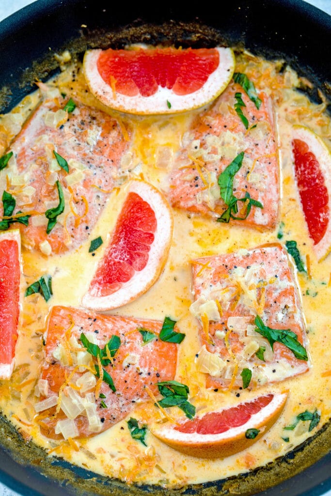 Overhead view of a skillet of creamy grapefruit salmon with four salmon filets, basil, onions, and grapefruit wedges in a grapefruit cream sauce.