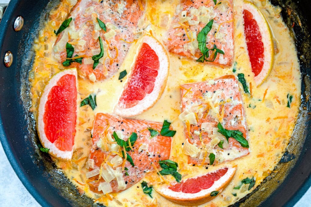 Landscape view of a skillet with creamy grapefruit salmon, including salmon filets, grapefruit wedges, chopped onions, basil, and a grapefruit cream sauce.