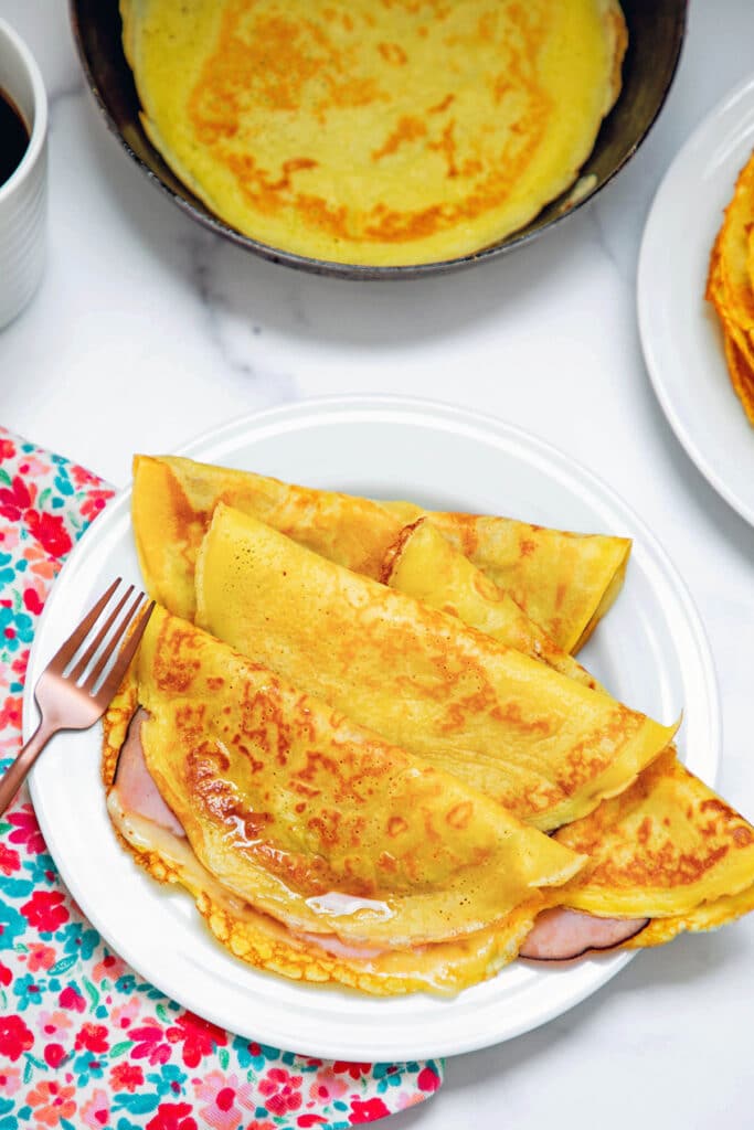 Three ham and cheese crepes with pancake mix folded over on plate with fork with crepe pan in background