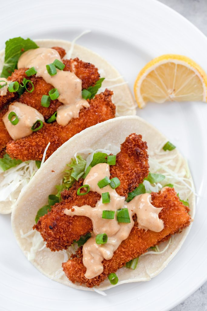 Overhead closeup of two fried fish tacos topped with hoisin mayo on white plate with lemon wedge on the side