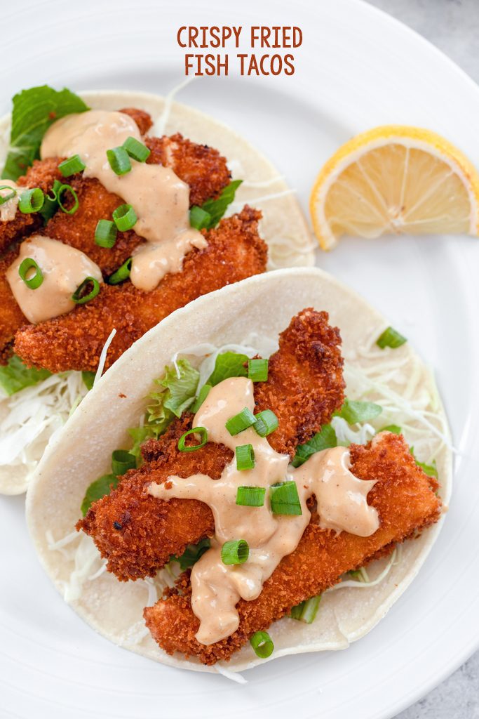 Overhead view of a white plate with two fried fish tacos with hoisin mayo and scallions with a lemon wedge on the side and recipe title at top