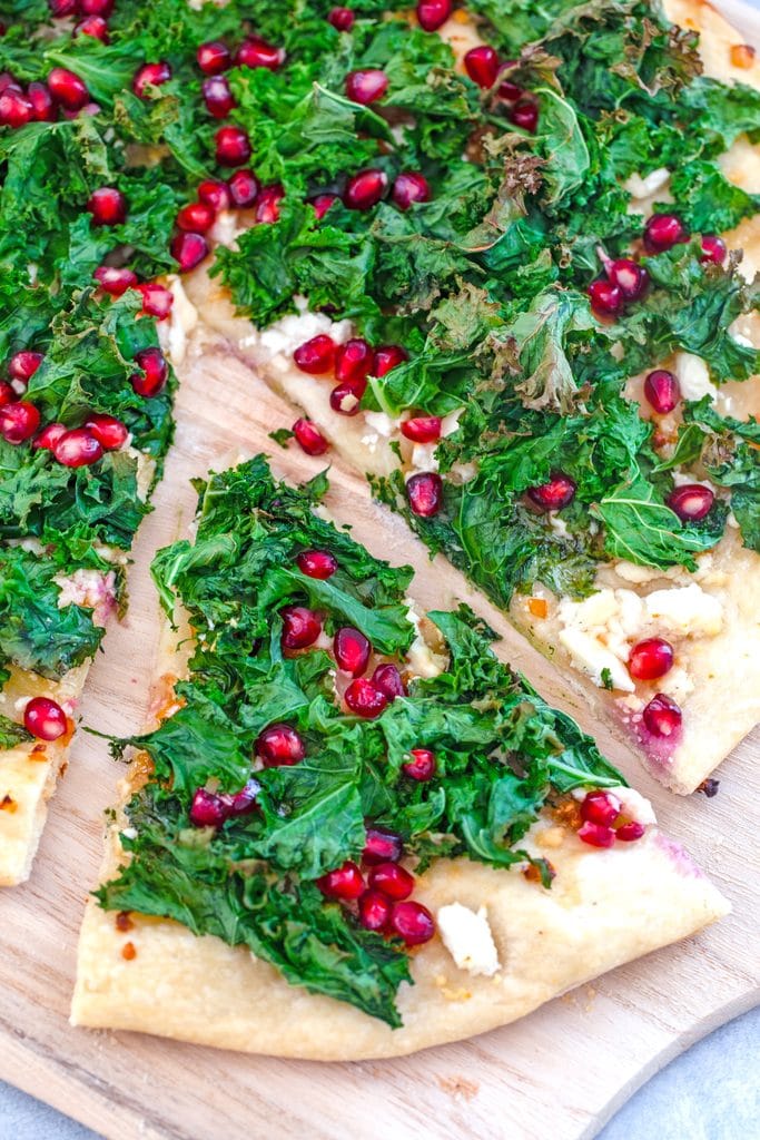 Overhead view of kale flatbread topped with crispy kale, feta, and pomegranate arils with a slice pulled out from rest of pizza