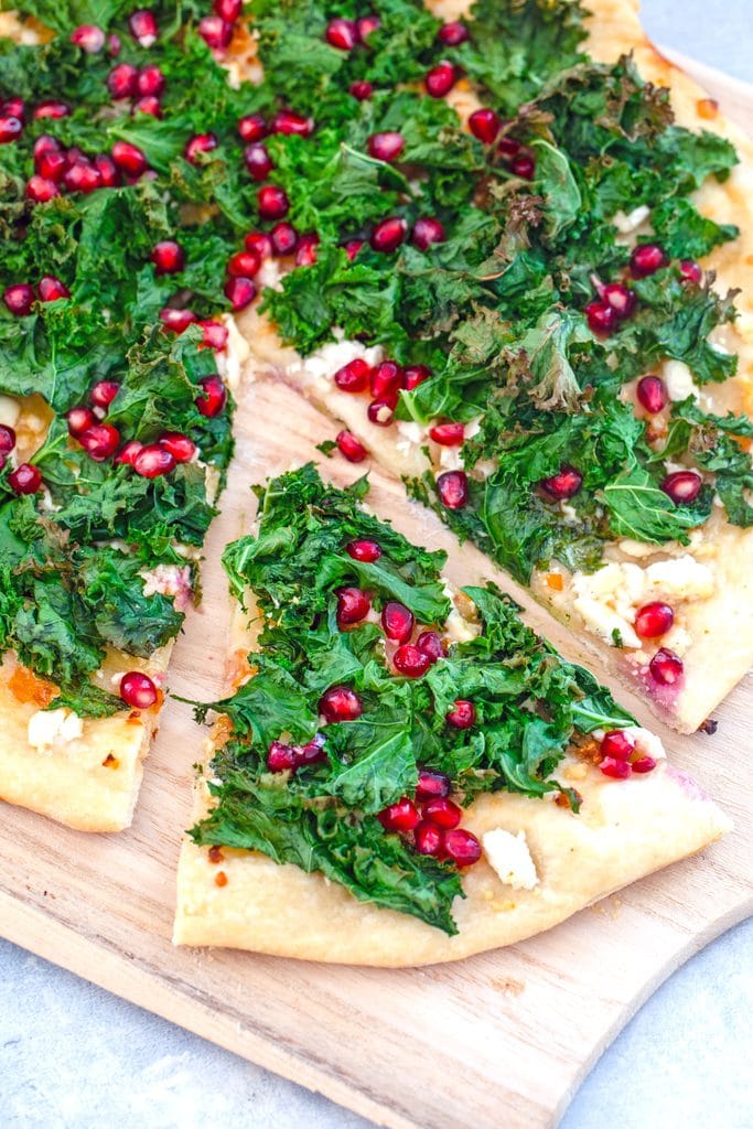 Overhead view of kale flatbread pizza topped with kale, feta, and pomegranate on a wooden board with one slice pulled out