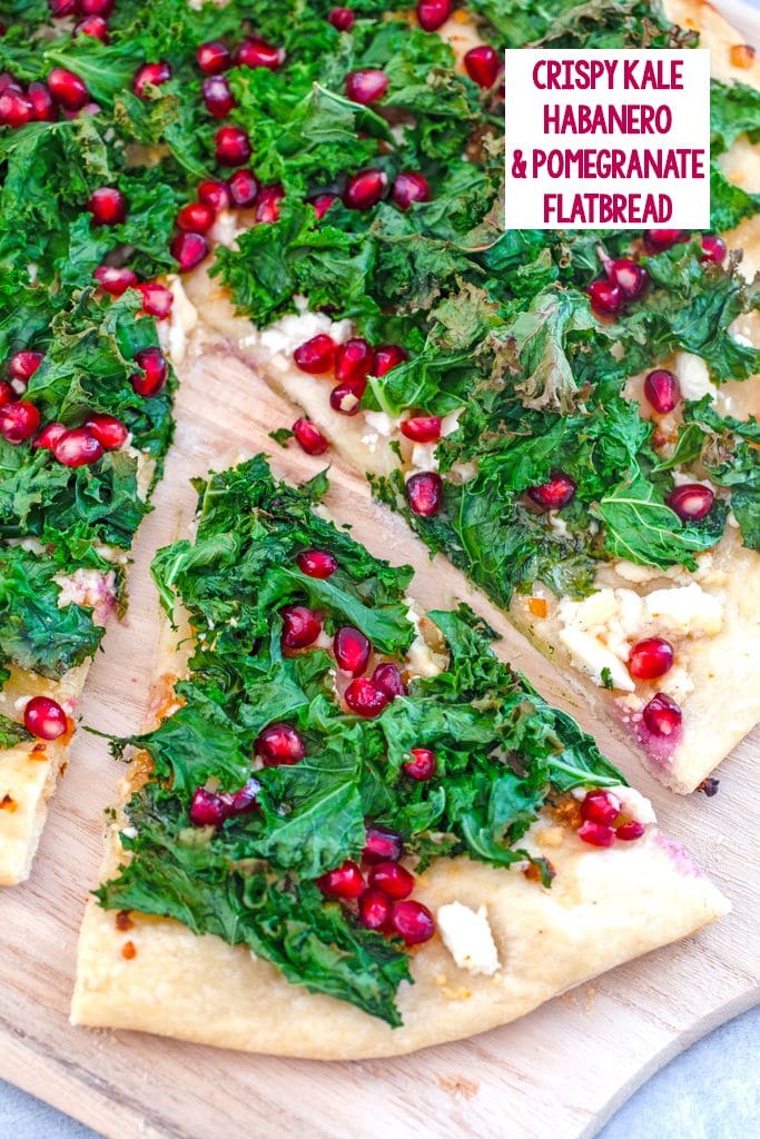 Overhead view of kale flatbread topped with crispy kale, feta, and pomegranate arils with a slice pulled out from rest of pizza and recipe title at top