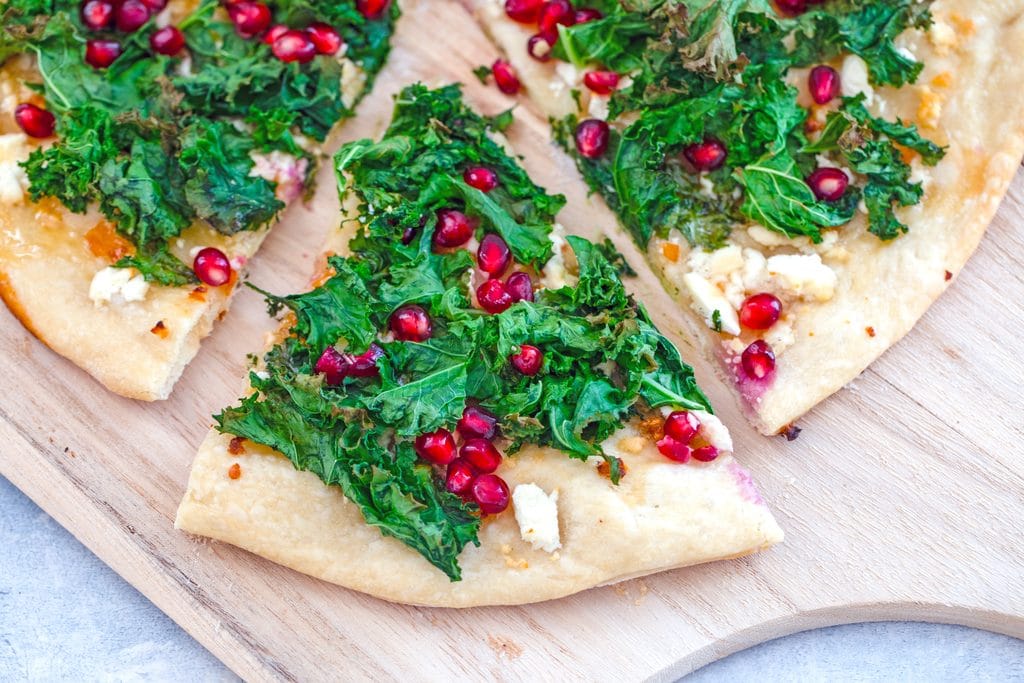 Landscape closeup image of slice of kale flatbread pizza with pomegranate and habanero being pulled out from rest of pizza on wooden board