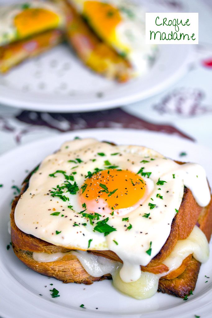 Head-on view of a croque madame with egg, mornay sauce, and parsley on a white plate with a second sandwich in the background and recipe title at top