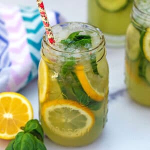 View of a mason jar filled with cucumber lemonade with sliced lemons and basil in it and more glasses of lemonade in background