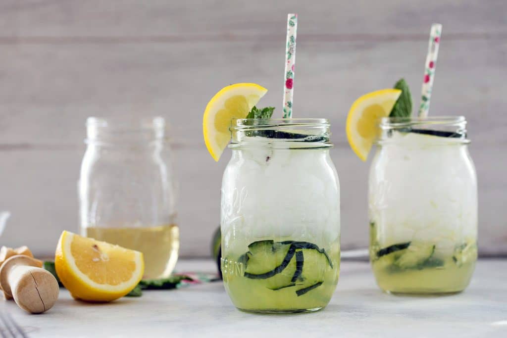 View of two mason jars with cucumber collins with muddled cucumbers in the bottom and topped with lemon wedge, cucumber slice, mint, and floral straw. Lemon wedge and jar of simple syrup in the background