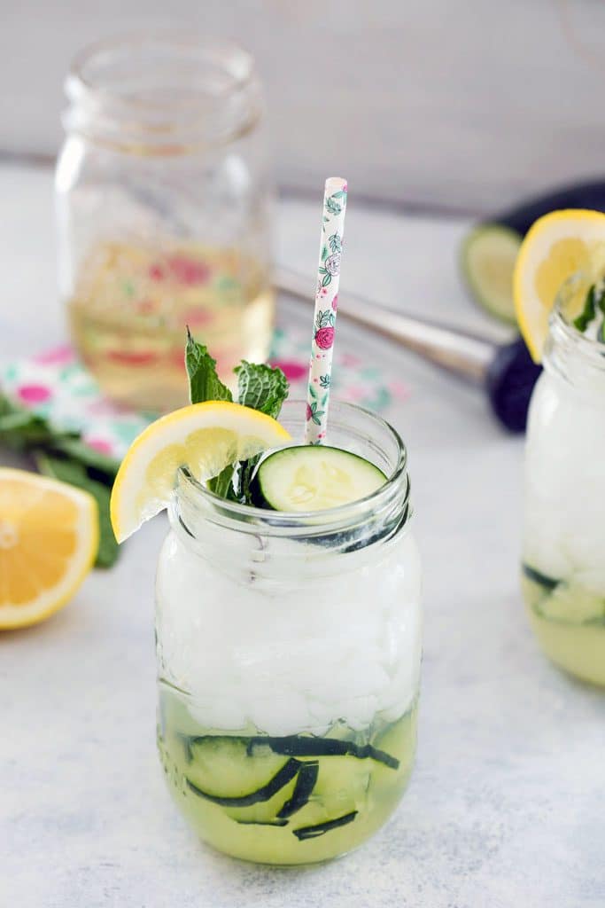 Cucumber collins cocktail with muddled cucumber in the glass and cucumber slice, lemon, mint, and floral straw as garnish. Jar of simple syrup, lemon, mint, cucumber, and muddler in the background