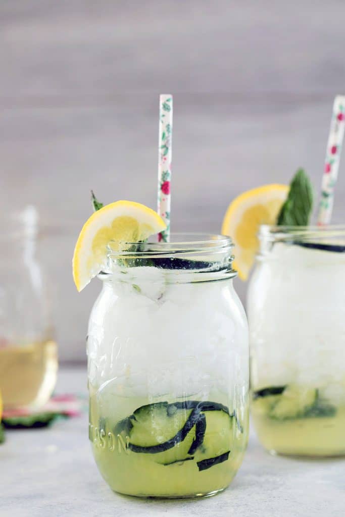 Head-on view of mason jar filled with cucumber collins with cucumbers muddled at the bottom of the glass and topped with a lemon wedge and floral straw and jar of simple syrup in the background