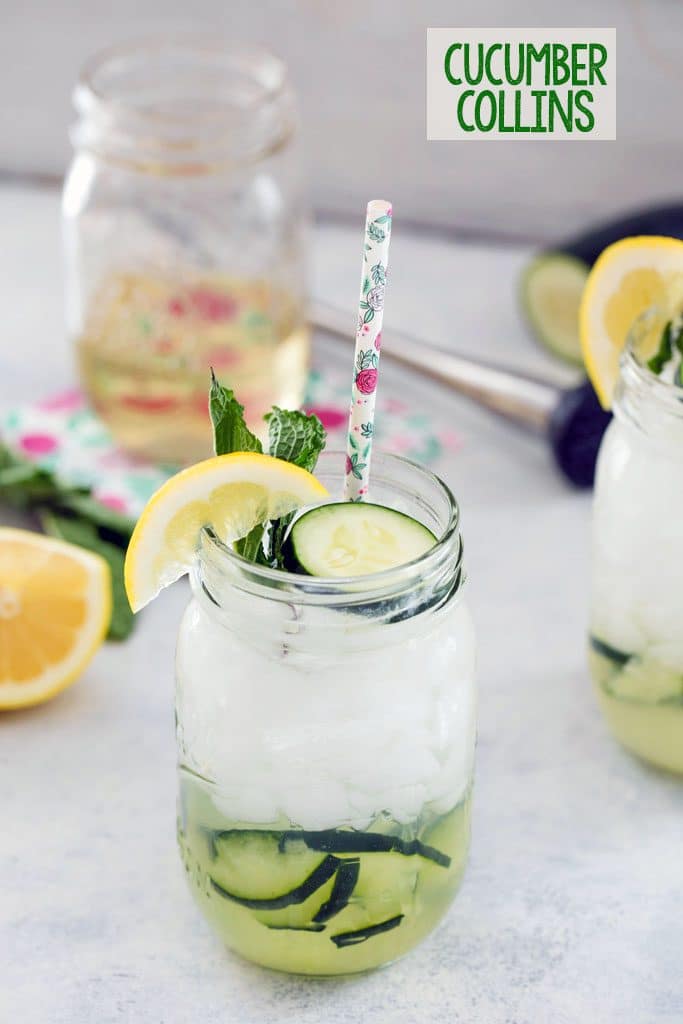 Cucumber collins cocktail with muddled cucumber in the glass and cucumber slice, lemon, mint, and floral straw as garnish. Jar of simple syrup, lemon, mint, cucumber, and muddler in the background and "Cucumber Collins" text at the top