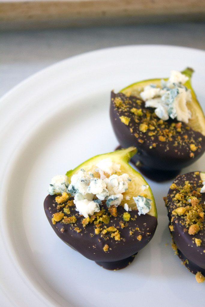 Close-up of three chocolate figs on a white plate topped with gorgonzola cheese and crushed pistachios