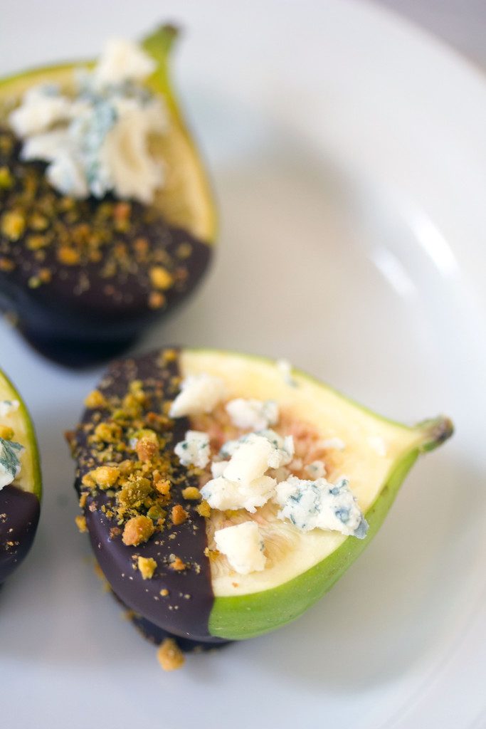 A close-up of a dark chocolate-dipped fig topped with gorgonzola cheese and crushed pistachios on a white plate with "Dark Chocolate-Dipped Figs with Gorgonzola" text on top