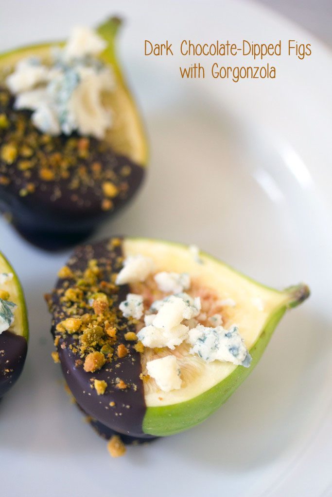 A close-up of a dark chocolate-dipped fig topped with gorgonzola cheese and crushed pistachios on a white plate with "Dark Chocolate-Dipped Figs with Gorgonzola" text on top