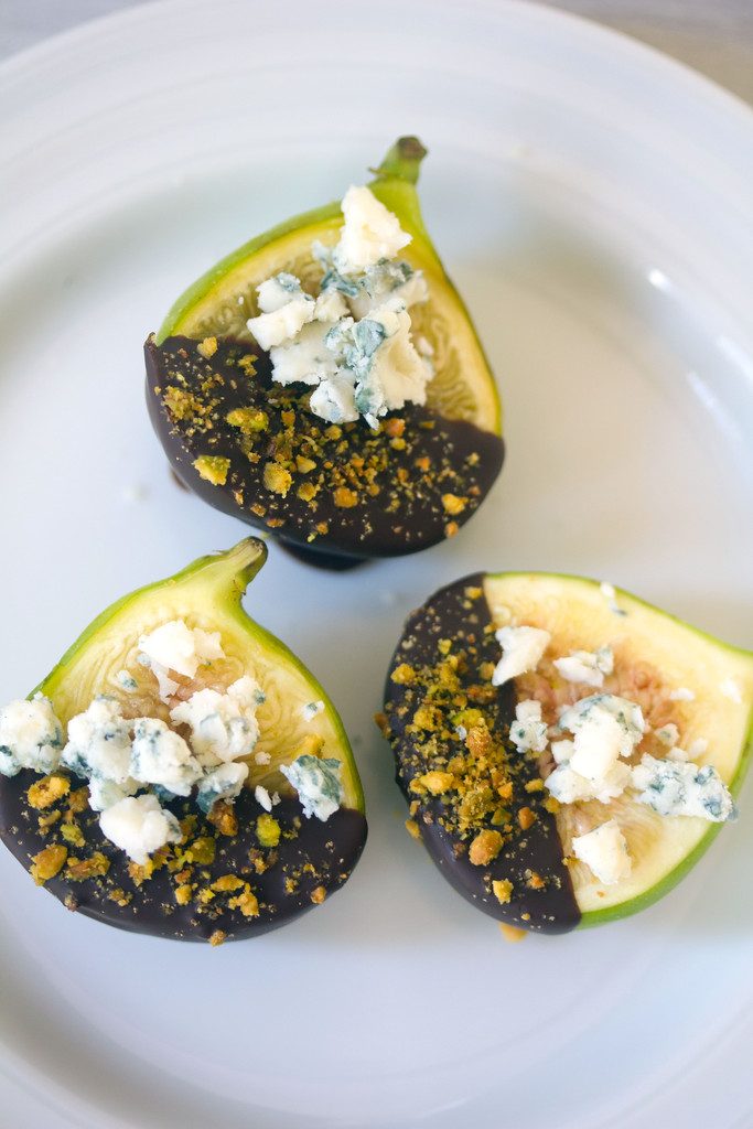 Bird's eye vier of three chocolate dipped figs topped with gorgonzola cheese and crushed pistachios on a white plate