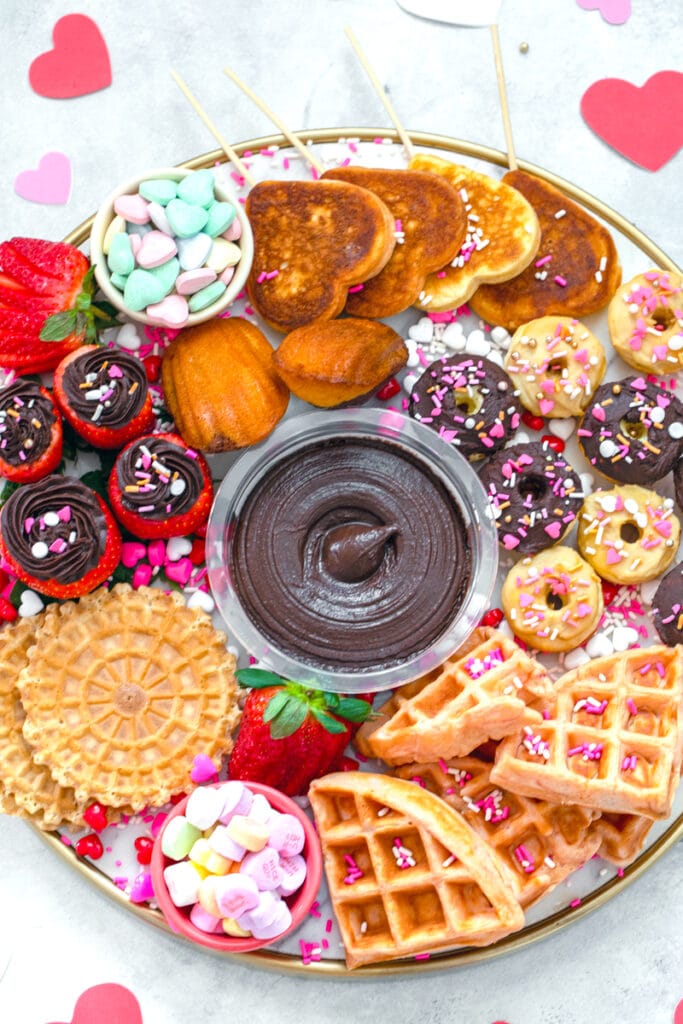 overhead view of a dessert hummus brunch board featuring dark chocolate hummus surrounded by mini donuts and waffles, heart-shaped pancakes, hummus filled strawberries, pizzells, madeleines, and Valentine's Day candy