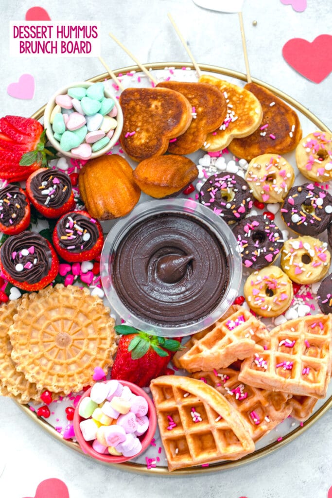 overhead view of a dessert hummus brunch board featuring dark chocolate hummus surrounded by mini donuts and waffles, heart-shaped pancakes, hummus filled strawberries, pizzells, madeleines, and Valentine's Day candy with "dessert hummus brunch board" text at top
