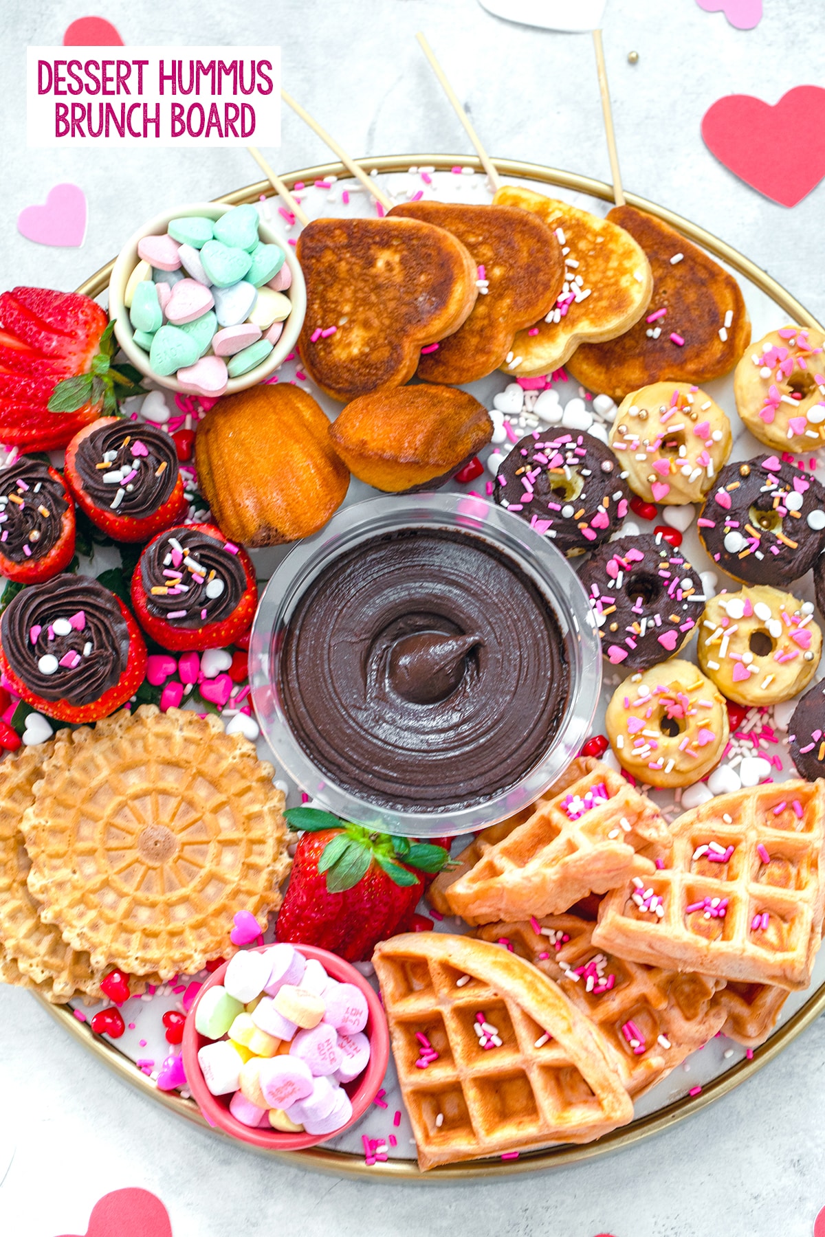 Overhead view of a dessert hummus brunch board featuring dark chocolate hummus surrounded by mini donuts and waffles, heart-shaped pancakes, hummus filled strawberries, pizzells, madeleines, and Valentine's Day candy with "dessert hummus brunch board" text at top.