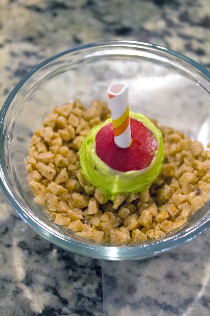 Overhead view of mini caramel candy apple coated in candy melts and being dipped into heath bar crumbles