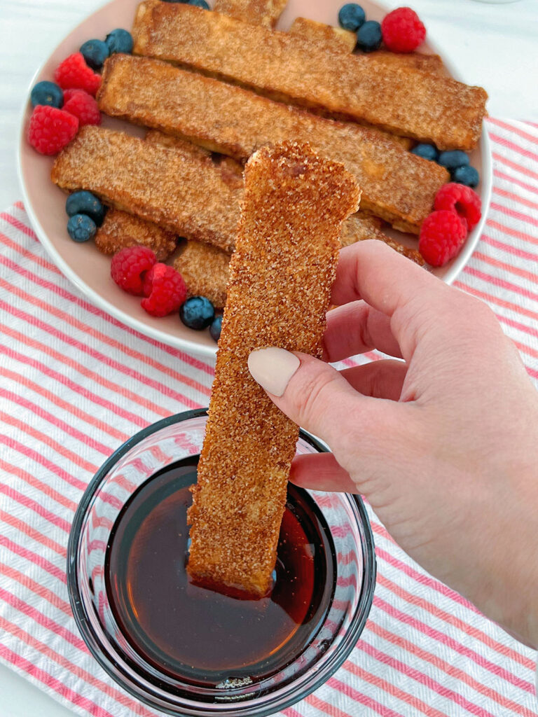 Dipping naan french toast stick into maple syrup with more french toast sticks in background.