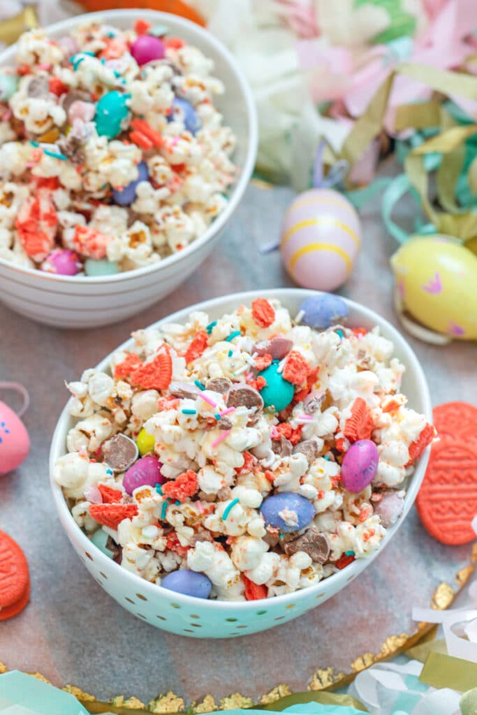 View of a bowl of Easter candy popcorn with M&Ms, sprinkles, and Easter Oreo cookies with second bowl and Easter eggs in background