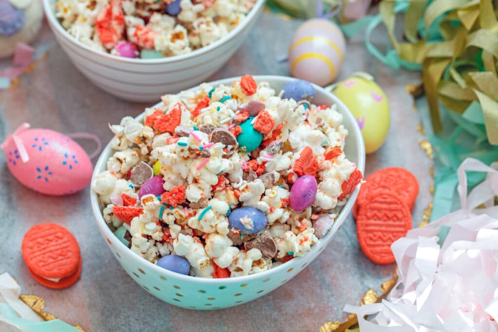 Landscape view of a bowl of Easter candy popcorn with M&Ms and sprinkles surrounded by Easter eggs and Oreo cookies with a second bowl of popcorn in background