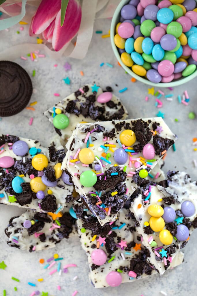 Bird's eye view of stack of white chocolate Easter Oreo bark with spring M&ms, Peeps Oreo cookies, and pastel sprinkles with bowl of M&Ms, pink tulips, cookies, and sprinkles in the background.