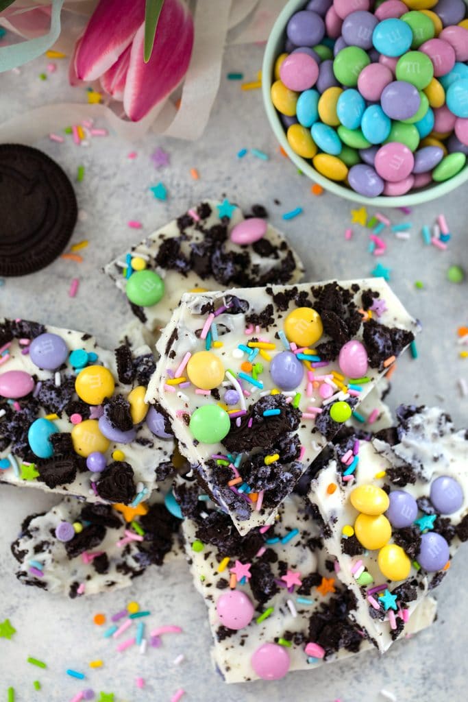 Bird's eye view of stack of white chocolate Easter Oreo bark with spring M&ms, Peeps Oreo cookies, and pastel sprinkles with bowl of M&Ms, pink tulips, cookies, and sprinkles in the background
