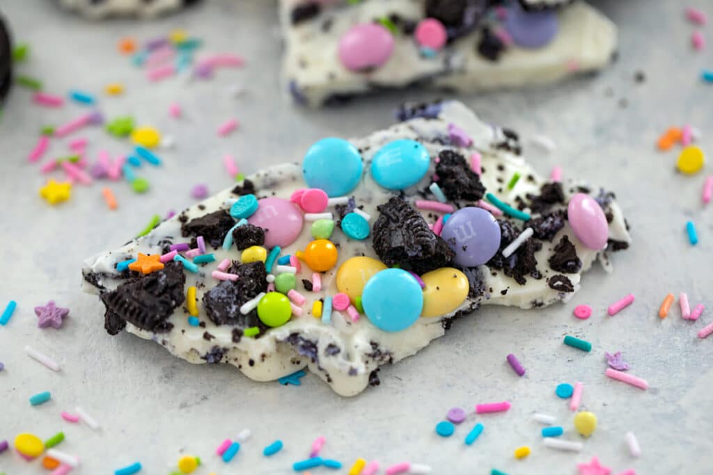 Landscape view of a piece of white chocolate Easter Oreo bark with spring M&Ms, crushed Peeps Oreo cookies, and pastel sprinkles with sprinkles scattered all around.