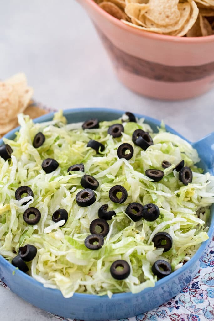 Closeup head-on view of easy taco dip topped with shredded lettuce and sliced olives with bowl of chips in background.