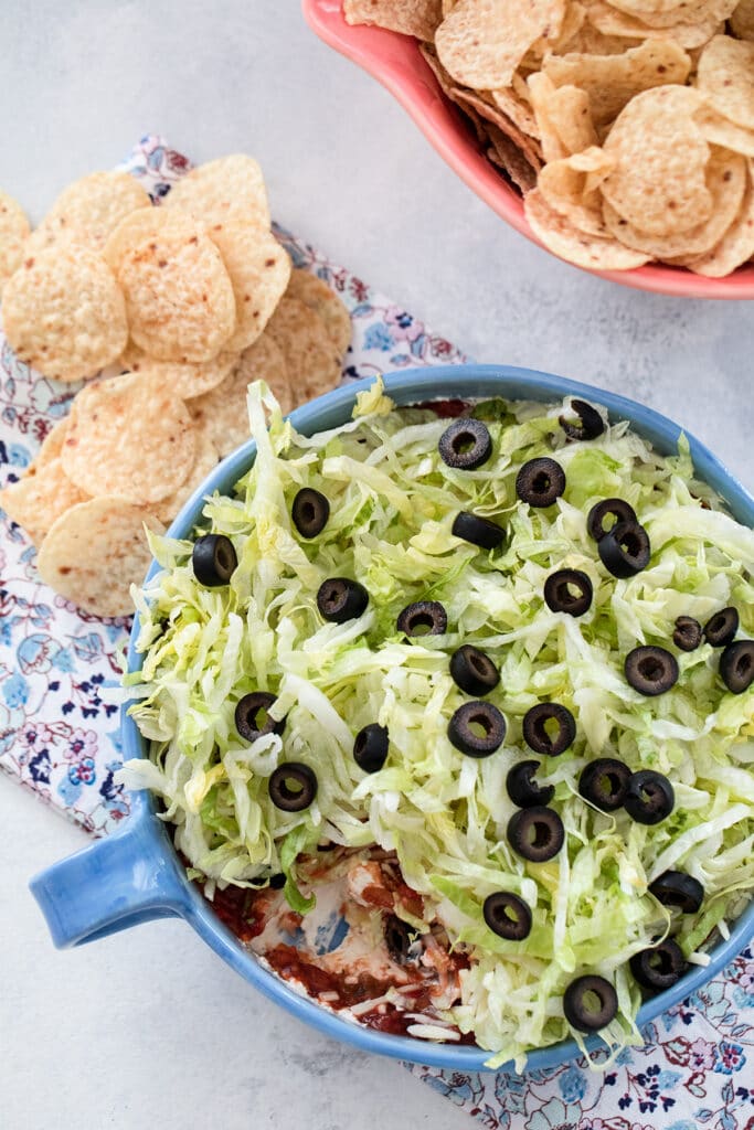 Overhead view of a taco dip with cream cheese, salsa, cheese, lettuce, and olives with chips all around.