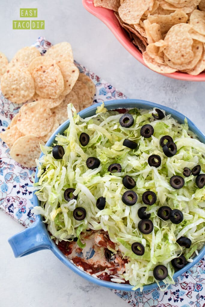 Overhead view of a taco dip with cream cheese, salsa, cheese, lettuce, and olives with chips all around with recipe title at top.