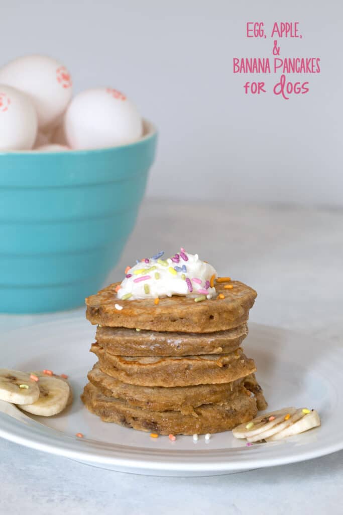 Stack of egg, apple, and banana pancakes for dogs with whipped cream and sprinkles and bowl of eggs in the background with recipe title at top