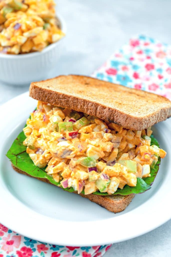 Head-on view of a sandwich with egg salad with lots of crunch with Swiss chard and with a bowl of more crunchy egg salad in the background