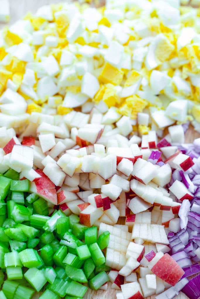 Overhead close-up view of chopped hard boiled eggs, chopped apples, chopped celery, and chopped red onion on cutting board