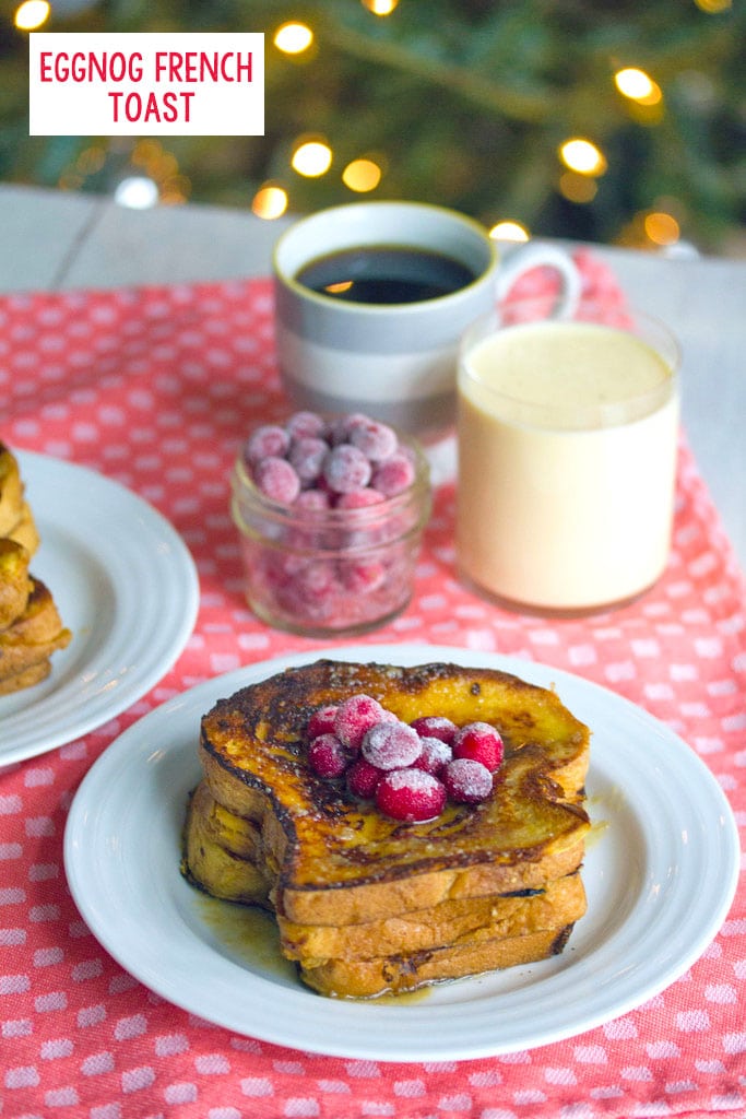 Head-on view of a plate of eggnog french toast topped with sugared cranberries with cup of cranberries, eggnog, and coffee in the background and recipe title at top