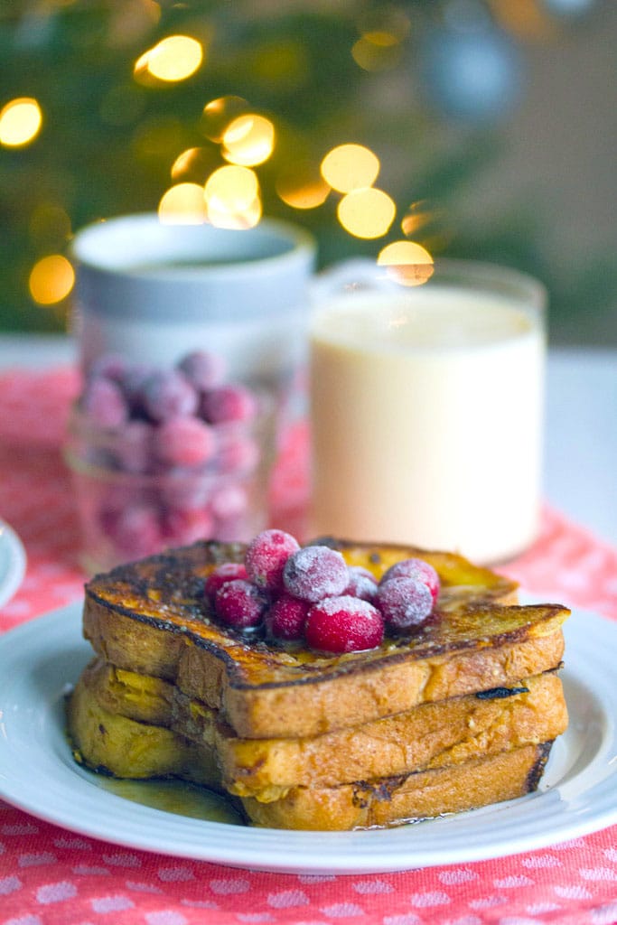 Head-on view of a stack of eggnog french toast topped with sugared cranberries with jar of cranberries, glass of eggnog, and mug of coffee in background
