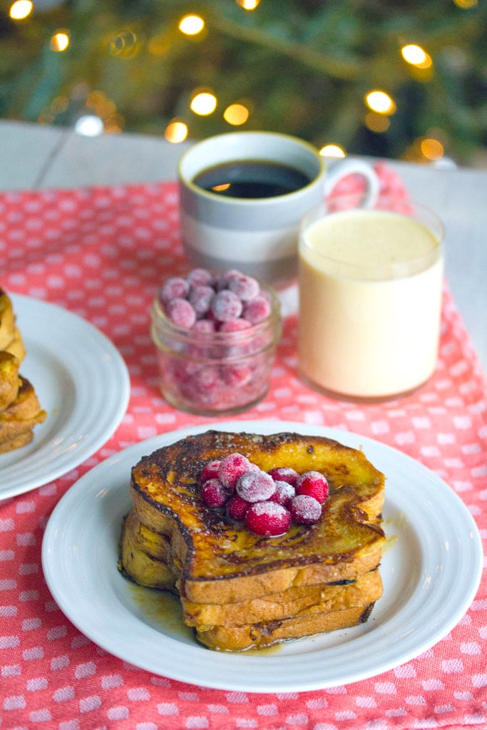 Head-on view of a plate of eggnog french toast topped with sugared cranberries with cup of cranberries, eggnog, and coffee in the background