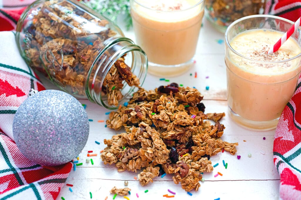 Landscape head-on view of eggnog granola spilling out of mason jar with two glasses of eggnog and silver ornament in background