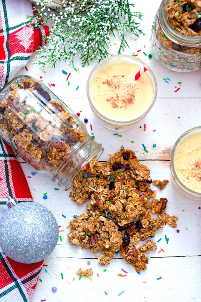 Bird's eye view of eggnog granola spilling out of mason jar with two glasses of eggnog, sprinkles, an ornament, and second jar of granola in background
