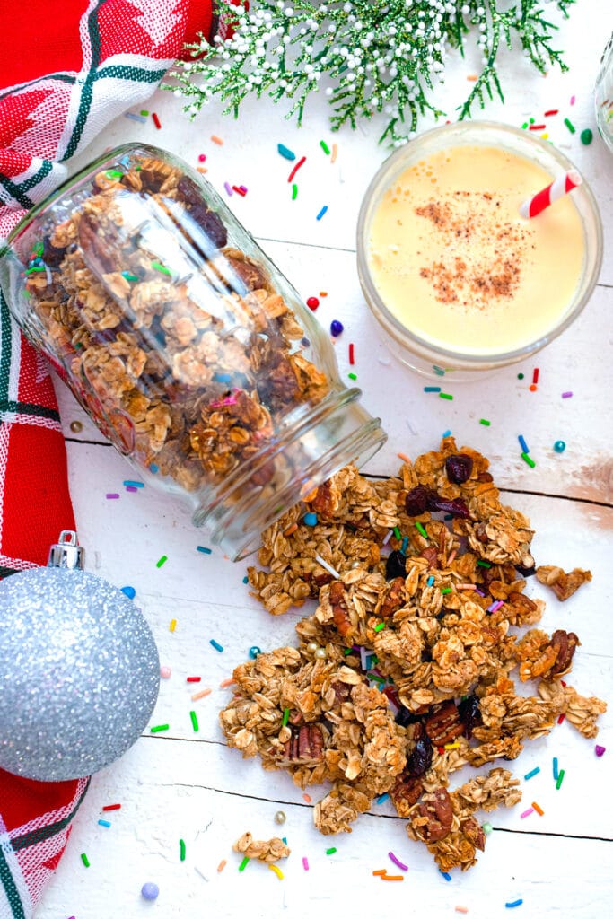 Overhead view of mason jar with eggnog granola spilling out of it with glass of eggnog, silver ornament, and sprinkles in background