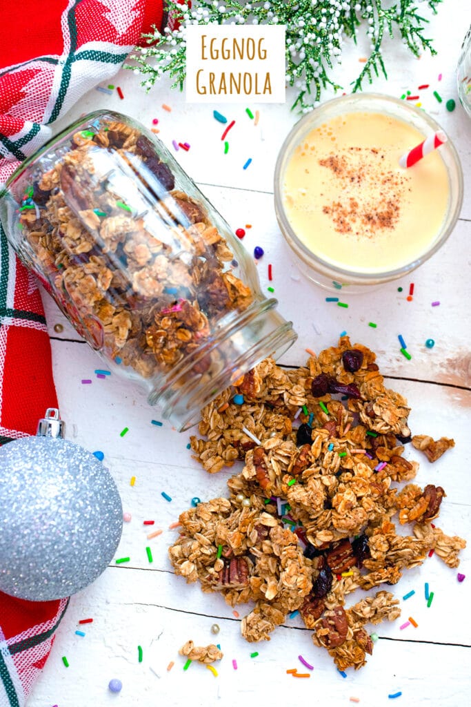 Overhead view of mason jar with eggnog granola spilling out of it with glass of eggnog, silver ornament, and sprinkles in background with recipe title at top